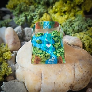 May include: A blue and green resin keycap with a small blue creature inside. The keycap is shaped like a cube and has a clear resin top.
