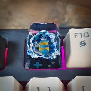 May include: A blue and yellow Pokemon character is encased in clear resin with white and blue swirls. The resin is set in a keycap for a mechanical keyboard.