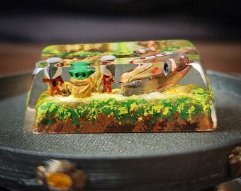 May include: A clear resin keycap with a miniature scene of a spaceship and three figures from the Star Wars franchise. The figures are painted in green, brown, and orange. The spaceship is painted in brown and white.