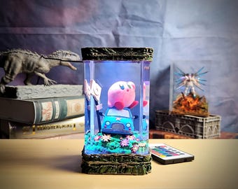 May include: A decorative, rectangular light-up display case featuring a pink character atop a blue car. The case has a detailed, textured base and top, with a colorful interior scene. A remote control sits nearby.