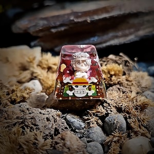 May include: A clear resin keycap featuring a detailed illustration of a character from the anime One Piece. The character is wearing a red and white hat and a white shirt. The background of the keycap is a red and gold design with a skull and crossbones.