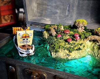 以下が含まれることがあります： 緑と青のレジン製の海を表現したミニチュアの宝箱。骸骨と交差した骨と麦わら帽子を被った小さな玩具の船が海を航海しています。シーンは緑の苔と赤いキノコのある小さな島で囲まれています。