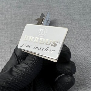 May include: A silver rectangular money clip with the word "BRABUS" in capital letters and the words "fine leather" in a cursive font. The clip has a small metal hinge and is held by a gloved hand.