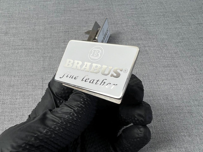May include: A silver rectangular money clip with the word "BRABUS" in capital letters and the words "fine leather" in a cursive font. The clip has a small metal hinge and is held by a gloved hand.