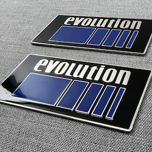 Peut inclure: Deux emblèmes rectangulaires noirs avec le mot "evolution" en lettres blanches. Chaque emblème présente un motif rectangulaire bleu avec trois bandes bleues verticales. Les emblèmes ont une bordure argentée.