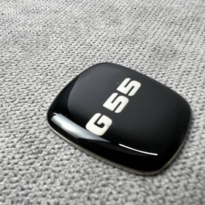Könnte beinhalten: Ein schwarzes, glänzendes Emblem mit dem Text "G55" in Weiß. Das Emblem hat eine abgerundete rechteckige Form und befindet sich auf einem grau strukturierten Hintergrund.