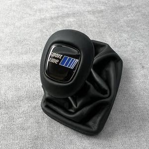 Könnte beinhalten: Ein schwarzer Schaltknauf aus Leder mit passendem Schaltsack aus schwarzem Leder. Die Oberseite des Knaufs zeigt ein glänzend schwarzes Emblem mit dem weißen Schriftzug Sport Line und vier blauen vertikalen Streifen im Verlauf.