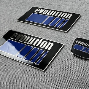 以下が含まれることがあります： 青いアクセントが付いた、黒とシルバーのEvolutionエンブレムが3つ。2つは長方形で、1つは楕円形です。「evolution」の文字はシルバーです。エンブレムは光沢のある仕上がりです。
