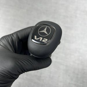 May include: Black shift knob with a silver emblem featuring a three-pointed star inside a circle. The text "V12" is below the emblem. The shift knob is held by a gloved hand.