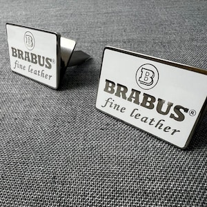 May include: Two silver cufflinks with a rectangular face. The face of each cufflink is white with the word "BRABUS" in a bold, sans-serif font, and "fine leather" below it. A circular logo is at the top.