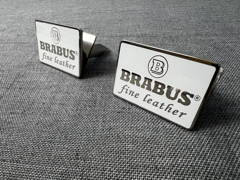 May include: Two silver cufflinks with a rectangular face. The face of each cufflink is white with the word "BRABUS" in a bold, sans-serif font, and "fine leather" below it. A circular logo is at the top.