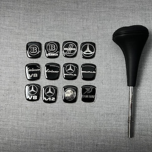 Puede incluir: Un conjunto de insignias negras para automóviles con logotipos blancos, como Brabus y Lorinser, junto con un pomo de palanca de cambios negro. Las insignias son rectangulares con esquinas redondeadas, y la palanca tiene un eje cromado.