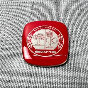 Puede incluir: Un emblema cuadrado rojo con esquinas redondeadas que presenta un escudo circular plateado. El escudo muestra un árbol a la izquierda, un resorte de válvula a la derecha y el texto AFFALTERBACH arriba. La palabra AMG está impresa en plata.