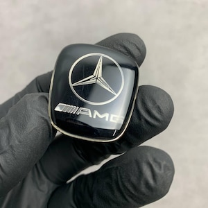 Op de afbeelding: Een zwart en zilver Mercedes-Benz embleem met een driepuntige ster-logo en de tekst "AMG" eronder.