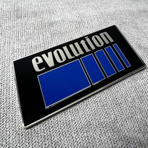 Puede incluir: Un emblema rectangular de metal negro con la palabra "evolution" en letras plateadas. El emblema presenta un diseño rectangular azul con bordes plateados. El emblema está sobre una superficie gris texturizada.