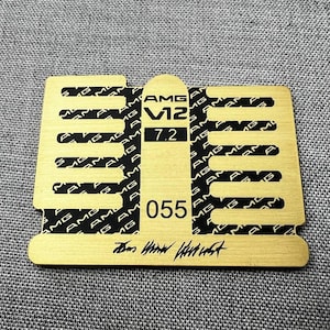 May include: A gold-colored rectangular emblem with black text and a unique shape. The emblem features the text "AMG V12 7.2 055" and the AMG logo repeated in a pattern. The bottom has a signature.