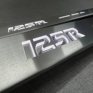Peut inclure: Deux protections de seuil de porte de voiture en métal brossé avec le texte "125R" en lettres blanches éclairées. Les protections sont rectangulaires et présentent un design élégant et moderne. Le fond est une surface gris foncé.