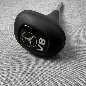 May include: Black gear shift knob with a silver shaft. The knob features a silver Mercedes-Benz logo and the letters "V8" in silver. The knob is made of black leather or a similar material.