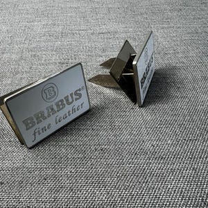 May include: Two silver car air vent clips with the text "BRABUS fine leather" in a gray font. The clips are rectangular with a silver metal finish and a clip on the back. The background is a gray fabric.