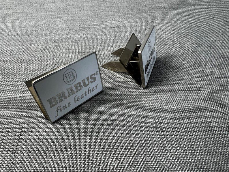 May include: Two silver car air vent clips with the text "BRABUS fine leather" in a gray font. The clips are rectangular with a silver metal finish and a clip on the back. The background is a gray fabric.