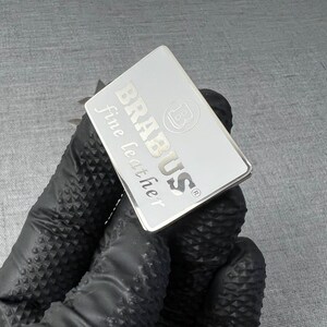 May include: A rectangular silver tag with the word "BRABUS" and "fine leather" printed on it. The tag also features the "B" logo. The tag is held by a person wearing a black glove.