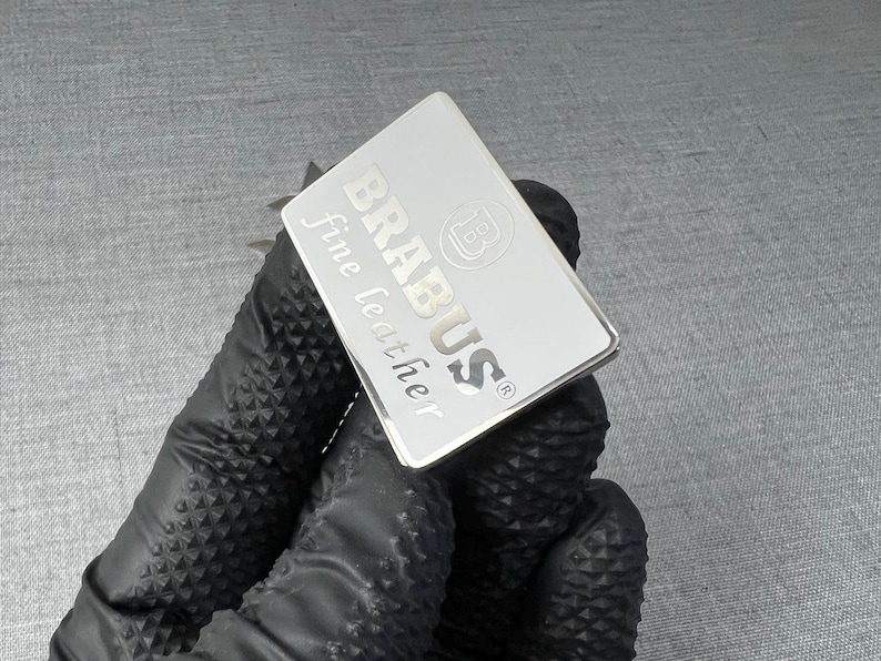 May include: A rectangular silver tag with the word "BRABUS" and "fine leather" printed on it. The tag also features the "B" logo. The tag is held by a person wearing a black glove.