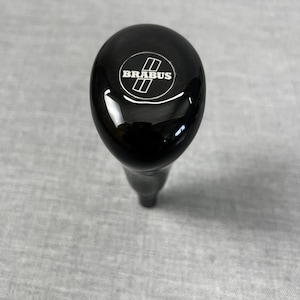 May include: A black gear shift knob with a glossy finish. The knob features the white "BRABUS" logo with two parallel rectangles. The gear shift is designed for a vehicle.