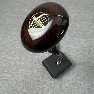 Könnte beinhalten: Dunkelbrauner, ovaler Schalthebelknauf mit goldfarbenem "BRABUS"-Schriftzug. Der Knauf ist auf einem schwarzen Sockel mit einer klaren, gebogenen Halterung montiert.