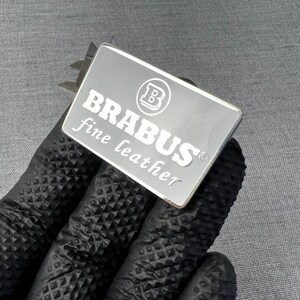May include: A rectangular silver metal tag with the word "BRABUS" in bold white letters and the words "fine leather" in a smaller font. The tag also features a circular logo. The tag is held by a person wearing a black glove.