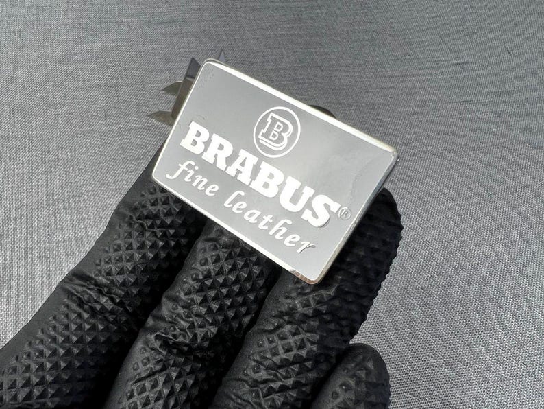 May include: A rectangular silver metal tag with the word "BRABUS" in bold white letters and the words "fine leather" in a smaller font. The tag also features a circular logo. The tag is held by a person wearing a black glove.