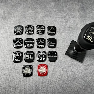 Könnte beinhalten: Eine Sammlung von vierzehn kleinen, quadratischen, glänzenden Auto-Emblemen mit verschiedenen Logos wie Brabus, AMG, V8, V12, Carlsson, Lorinser und Wald, neben einem schwarzen Schaltknauf mit einer Basis aus Kohlefaser.