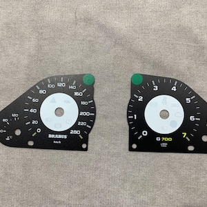 Può includere: Due indicatori del cruscotto dell'auto neri con quadranti bianchi. L'indicatore sinistro è etichettato "BRABUS" e ha un tachimetro che arriva a 280 km/h. L'indicatore destro è etichettato "G 700" e ha un contagiri che arriva a 7.
