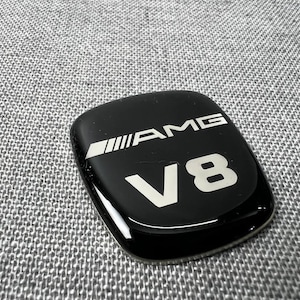 May include: A black, glossy AMG V8 emblem with white lettering. The emblem has a rectangular shape with rounded corners and a silver stripe design on the left side. The text "AMG" and "V8" are prominently displayed.