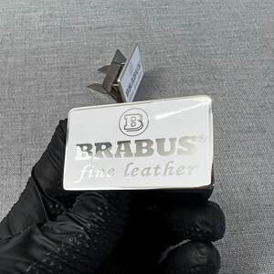 May include: A white rectangular product label with the word "BRABUS" in silver lettering and a logo. The words "fine leather" are below the brand name. The label is held by a gloved hand.