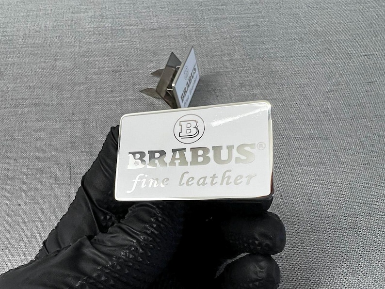 May include: A white rectangular product label with the word "BRABUS" in silver lettering and a logo. The words "fine leather" are below the brand name. The label is held by a gloved hand.