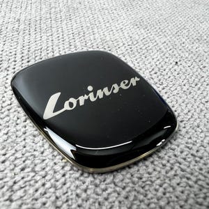 Könnte beinhalten: Ein schwarzes, rechteckiges Emblem mit abgerundeten Ecken. Das Wort "Lorinser" ist in einer kursiven, weißen Schrift über die Mitte geschrieben. Das Emblem hat eine glänzende, reflektierende Oberfläche und einen goldfarbenen Rand.