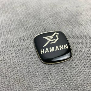 Peut inclure: Un emblème carré noir et argent avec le mot "HAMANN" en lettres blanches et un logo d'oiseau stylisé.