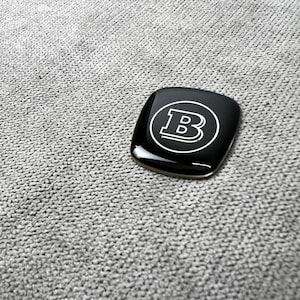 Peut inclure: Un emblème noir de forme carrée avec une finition brillante. Il présente la lettre blanche "B" à l'intérieur d'un cercle blanc. L'emblème est posé sur un fond de tissu gris texturé.