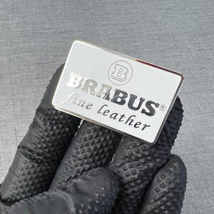 May include: A rectangular silver emblem with the word "BRABUS" in capital letters and "fine leather" in lowercase letters. The emblem has a silver border and a logo in the upper left corner. The emblem is held by a gloved hand.