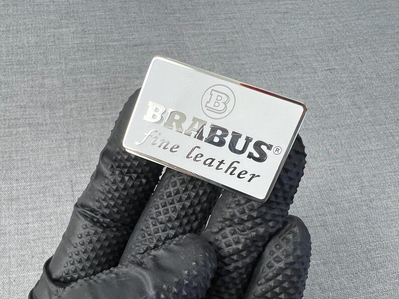 May include: A rectangular silver emblem with the word "BRABUS" in capital letters and "fine leather" in lowercase letters. The emblem has a silver border and a logo in the upper left corner. The emblem is held by a gloved hand.