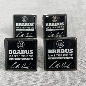 Può includere: Quattro emblemi quadrati neri con la scritta "BRABUS MASTERPIECE SUPERBLACK" in bianco e una firma. Ogni emblema ha un logo "B" argentato. Gli emblemi sono su una superficie grigia.
