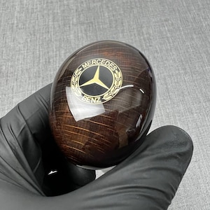 以下が含まれることがあります： 濃い茶色の木製メルセデス・ベンツのシフトノブ。ノブは光沢仕上げで、金色のメルセデス・ベンツのロゴが特徴です。ロゴはノブの上部に配置されています。手袋をはめた手で持っています。