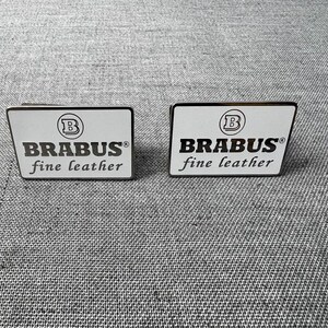 May include: Two rectangular, white and black Brabus fine leather badges. Each badge has the Brabus logo, a stylized "B" inside a circle, and the words "BRABUS fine leather" in a black, sans-serif font.