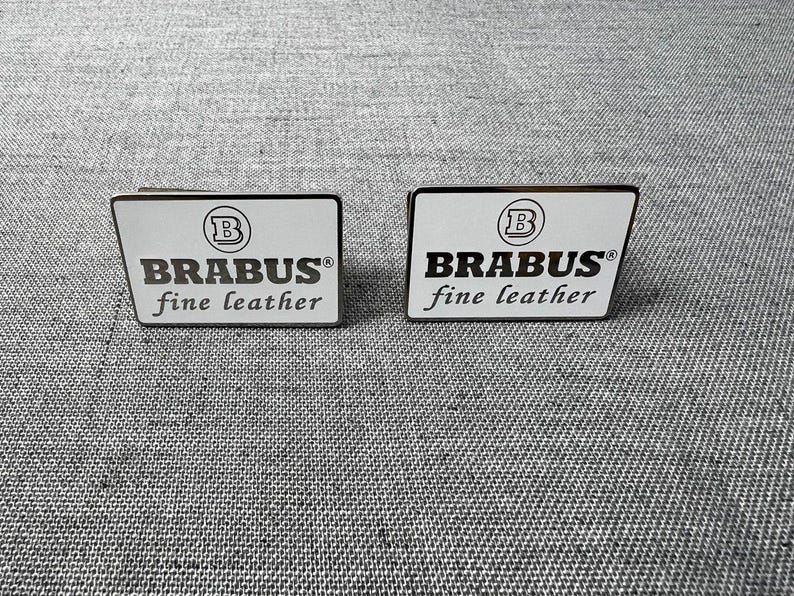 May include: Two rectangular, white and black Brabus fine leather badges. Each badge has the Brabus logo, a stylized "B" inside a circle, and the words "BRABUS fine leather" in a black, sans-serif font.