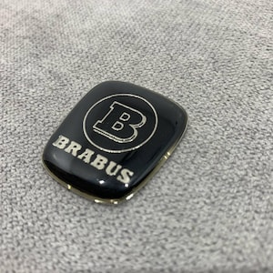 Può includere: Emblema di auto nero e argento con la parola "BRABUS" e una lettera "B" stilizzata all'interno di un cerchio.