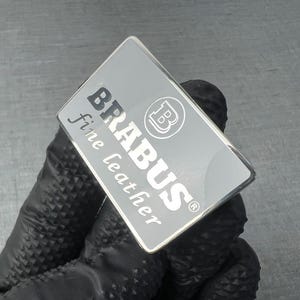 May include: A rectangular silver emblem with the word "BRABUS" in bold, black letters and "fine leather" in a smaller, cursive font. The emblem also features a circular logo with a "B" inside. The emblem is held by a gloved hand.
