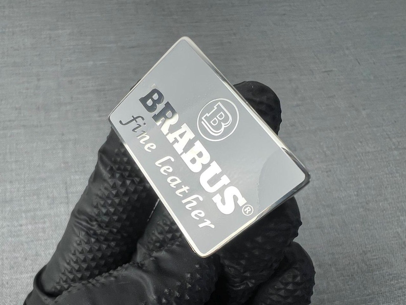 May include: A rectangular silver emblem with the word "BRABUS" in bold, black letters and "fine leather" in a smaller, cursive font. The emblem also features a circular logo with a "B" inside. The emblem is held by a gloved hand.