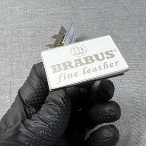 May include: A rectangular, silver and white leather tag with the text "BRABUS fine leather" in silver lettering. The tag has a silver metal border and a silver metal clip. The tag is held by a gloved hand.