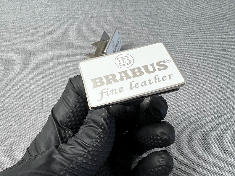 May include: A rectangular, silver and white leather tag with the text "BRABUS fine leather" in silver lettering. The tag has a silver metal border and a silver metal clip. The tag is held by a gloved hand.