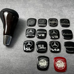 Op de afbeelding: Een zwarte versnellingspookknop van donker hout naast veertien kleine, rechthoekige, glanzende emblemen. De emblemen bevatten diverse autologo's en teksten, waaronder Brabus, AMG, V8, SL, V12, Carlsson, Lorinser, Wald en Affalterbach.